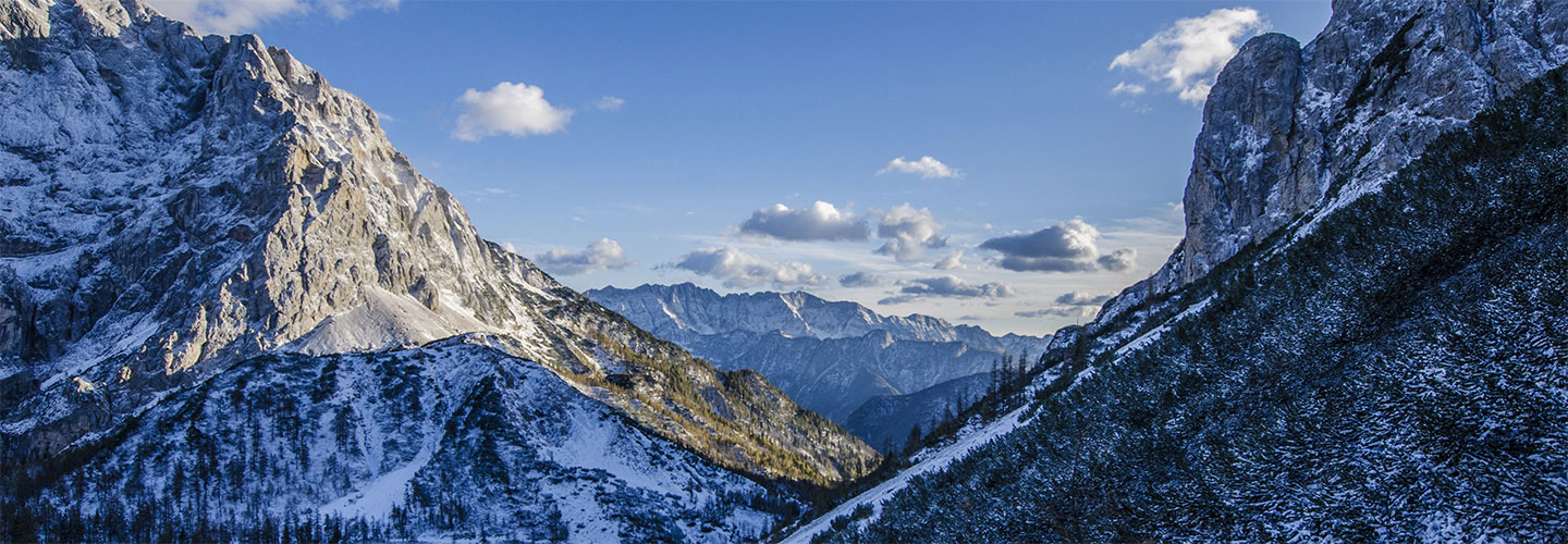 Panoramic view of snow-dusted rocky mountain peaks with evergreen trees under a blue sky with scattered white clouds
